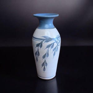 Può includere: Un vaso in ceramica con bordo azzurro e corpo bianco. Il vaso presenta un motivo floreale blu. Il vaso è alto circa 20 cm e ha un collo stretto.