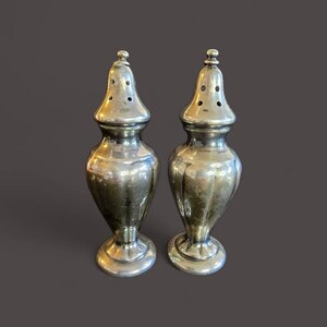 May include: A pair of antique silver salt and pepper shakers. Each shaker has a bulbous body, a flared base, and a conical top with several small holes. The shakers are made of a shiny, reflective metal.