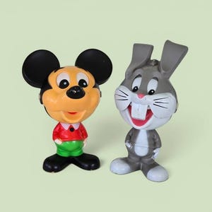 Pode incluir: Duas figuras de personagens em plástico. Uma é um rato com grandes orelhas pretas, rosto cor de pêssego, camisa vermelha e calças verdes. A outra é um coelho cinzento com orelhas compridas, bochechas brancas, dentes frontais e barriga cinzenta.
