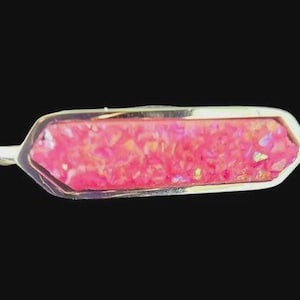 Può includere: Spilla rettangolare color oro con un vivace centro rosa e strutturato. La spilla ha una forma allungata unica ed è posta su uno sfondo nero. Il centro rosa presenta macchie dorate.