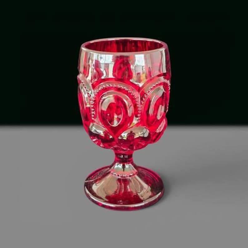 May include: A vibrant ruby red glass goblet featuring a prominent embossed pattern of repeating oval shapes and circular motifs. The vessel has a thick, sturdy stem and a wide, circular base, all crafted from translucent, deep red coloured glass.