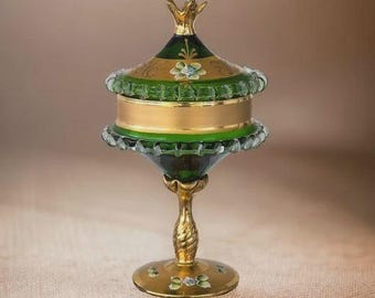 May include: An ornate, decorative glass container with a lid. The container features a gold base and accents, with green and clear glass elements. The lid is topped with a gold finial. The container is likely used for display or storage.