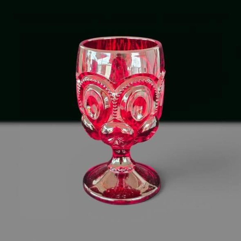May include: A vibrant ruby red glass goblet featuring a pressed pattern of circular motifs and starburst designs. The vessel has a thick stem and a wide, circular base, with a translucent finish that catches light across its textured surface.