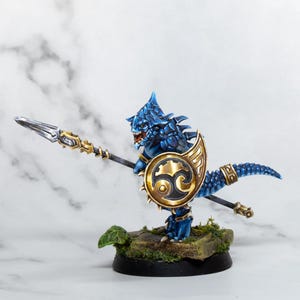 May include: A blue and gold miniature figurine of a lizard-like creature holding a spear and shield. The creature has a spiky back and a fierce expression. The shield is decorated with a gold and black design.