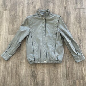 May include: A light gray jacket with a high collar and button closure. The jacket has long sleeves with buttoned cuffs and a ribbed hem. The material appears to be leather or a leather-like fabric. The jacket is laid flat on a wooden surface.