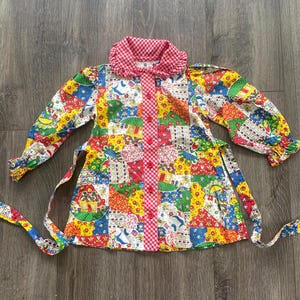 May include: A vibrant patchwork dress with a red gingham collar and long sleeves. The dress has a button-up front and a matching tie belt. The fabric is a mix of floral, animal, and geometric patterns in red, yellow, blue, and green.
