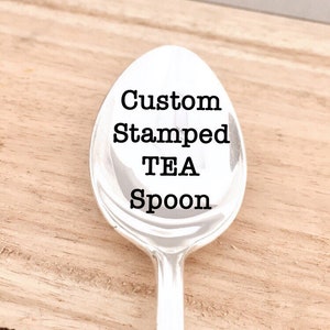 May include: A silver tea spoon with the words "Custom Stamped TEA Spoon" in black, centered on the spoon's bowl. The spoon is set against a light brown wooden surface.