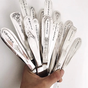 May include: A collection of vintage silver spoons, each engraved with the names of herbs and vegetables such as "Lettuce," "Basil," and "Rosemary." The spoons are held together, displaying their ornate designs and the clear, black lettering.
