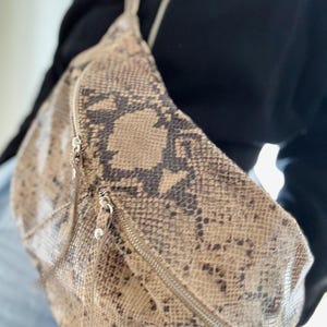 May include: A crossbody sling bag featuring a brown and black snakeskin pattern. The bag has a curved, rounded shape with two front-facing zippered compartments, each equipped with silver-toned metal zipper pulls and matching hardware.