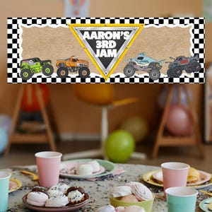 Puede incluir: Una pancarta de fiesta con un borde a cuadros blanco y negro y un fondo marr&oacute;n claro. La pancarta presenta cuatro monster trucks y el texto "AARON'S 3RD JAM" en un tri&aacute;ngulo amarillo. La mesa est&aacute; puesta con golosinas y vasos.