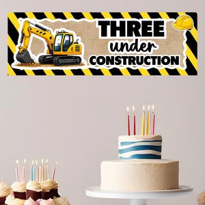 Peut inclure: Une bannière avec un dessin de pelle mécanique jaune et le texte "THREE under CONSTRUCTION". Un gâteau d'anniversaire avec des bougies et des petits gâteaux sont au premier plan. La bannière a une bordure rayée jaune et noire.