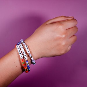May include: Three beaded bracelets with white letter beads spelling out names, interspersed with colourful beads. The bracelets are worn on a wrist against a purple background. The names are "ASUKA", "SHINJI", and "REI".