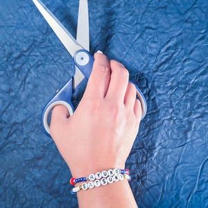 May include: Blue handled scissors with silver blades are held over a blue textured surface. Two beaded bracelets spell out "RYUKO" and "SATSUKI" in white letter beads, accented with red and blue beads.