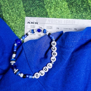 Puede incluir: Una pulsera de cuentas azules con las palabras "BLUE LOCK" deletreadas en cuentas de letras blancas. La pulsera está adornada con cuentas azules, negras y plateadas. Una camisa azul y una tarjeta con texto en el fondo.