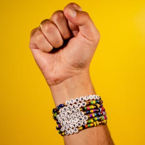 May include: A stack of beaded bracelets with white letter beads spelling out names like "JONATHON", "JOSEPH", and "JOTARO". The bracelets are in various colors, including blue, green, yellow, and red. The background is a solid yellow.