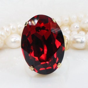 Puede incluir: Un anillo de gran tamaño con una piedra roja ovalada y detalles dorados. El anillo está sobre un fondo blanco.