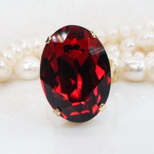 May include: A ring featuring a large, oval-cut, deep red faceted gemstone set in a gold-toned metal prong setting. The ring is positioned in front of a strand of white, round, lustrous pearls that are arranged in a soft, curved line behind it.