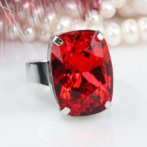 May include: A silver-toned ring featuring a large, rectangular, cushion-cut vibrant red gemstone held in place by four metal prongs. The ring band is smooth and metallic, showcasing a polished finish that reflects light across its surface.