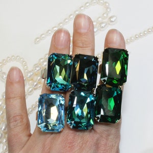 May include: A collection of five rectangular rings featuring large faceted crystals in shades of light blue, teal, and dark emerald green. Each ring is set in a metallic band, showcasing reflective surfaces and sharp geometric cuts.