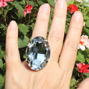 May include: A large oval-shaped faceted clear crystal ring set in a gold-toned metal prong setting. The crystal features a high-gloss finish with reflective surfaces that catch the light, creating a brilliant display of transparency and depth.
