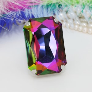 May include: A large, faceted, emerald-shaped, iridescent gemstone with a rainbow of colors, including red, green, blue, and purple. The gemstone is set in a silver-toned metal setting.
