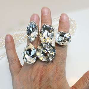 May include: Five rings featuring large, clear, faceted crystals in various shapes, including marquise, rectangular, round, and pear cuts, set in metallic bands. The crystals are highly reflective and clear, showcasing intricate geometric facets.