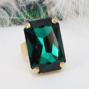 Può includere: Un grande anello con una pietra preziosa rettangolare verde smeraldo sfaccettata, montata su una fascia in metallo color oro lucido. La pietra presenta tagli geometrici netti che riflettono la luce, fissata da quattro piccole griffe dorate.