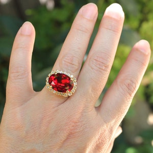 Könnte beinhalten: Ein goldener Ring mit einem großen, ovalen, roten Edelstein, der von kleineren, klaren Edelsteinen umgeben ist.