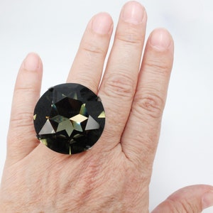 May include: A large, circular ring featuring a faceted, dark charcoal-gray stone with a star-shaped pattern cut into the center. The stone displays reflective, geometric surfaces that catch the light, creating a multi-dimensional appearance.