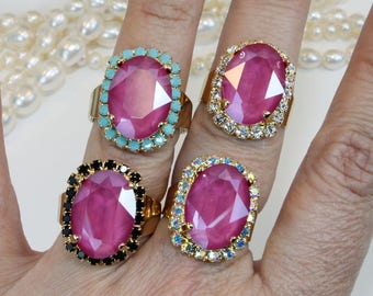May include: Four gold rings with large pink oval-shaped stones. Each ring has a different colored border of rhinestones. The rings are on a hand wearing a pearl necklace.
