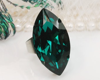 May include: A ring featuring a large, marquise-cut, deep emerald green faceted stone. The stone displays sharp geometric light reflections and is set on a simple, plain silver-toned metal band that supports the prominent, elongated gemstone.