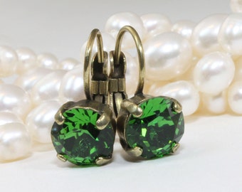 May include: A pair of leverback earrings featuring large, round, emerald green crystals set in antique brass-toned metal. The earrings are displayed against a backdrop of creamy white pearls, creating a classic and elegant look.
