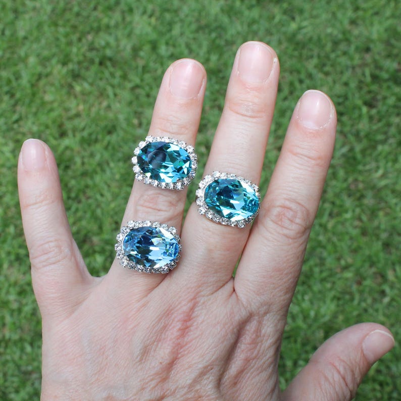 May include: Three silver-toned rings, each featuring a large, oval-cut, light blue gemstone surrounded by a halo of small, clear, sparkling stones. The rings are worn on the fingers, showcasing the faceted blue stones and the metallic settings.