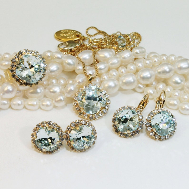May include: A set of gold jewellery with light blue crystal accents. The set includes a necklace, a ring, and a pair of earrings. The jewellery is displayed on a strand of white pearls.