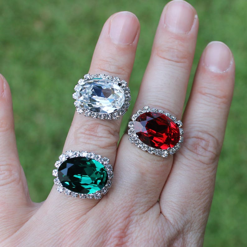 May include: Three oval-shaped rings featuring large faceted stones in clear, deep red, and emerald green. Each central stone is surrounded by a halo of small, clear, round-cut crystals set in a silver-toned metal band, creating a bold, reflective look.