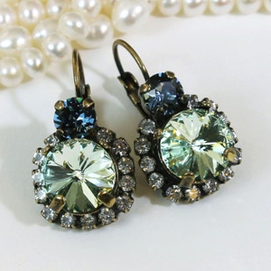 May include: A pair of dangle earrings with a light green crystal surrounded by smaller clear crystals and a smaller dark blue crystal on top. The earrings have a bronze finish.