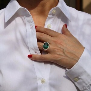 May include: A ring featuring a large, oval-cut dark green gemstone centered in a setting surrounded by a halo of small, clear, round-cut stones. The ring is worn on a finger, positioned against a crisp, white, button-down collared shirt.