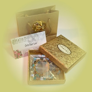 May include: A gold gift box with a turquoise and gold bracelet inside. The box is decorated with the words "HAND MADE TIMATIBO" and a website address. The box is sitting on a brown paper shopping bag with a gold rose decoration.