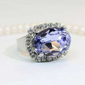 May include: A silver-toned ring featuring a large, oval-cut, light purple faceted stone as the centerpiece. The central stone is surrounded by a halo of small, clear, round-cut white crystals set in a silver-toned metal frame.