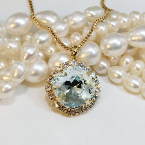 Puede incluir: Un collar de cadena dorada con un gran colgante redondo de cristal azul claro rodeado de cristales transparentes más pequeños. El collar se muestra sobre un lecho de perlas blancas.