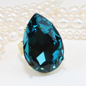 May include: A large, pear-shaped, deep teal faceted crystal ring set in a gold-toned metal band. The prominent, multi-faceted stone displays various shades of blue and green, reflecting light across its surface, positioned near white pearls.