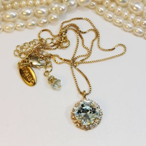 May include: A gold chain necklace featuring a large, round, clear crystal pendant encircled by smaller crystals. The pendant is secured to a gold chain with a lobster clasp. The chain also includes a small, clear crystal charm.