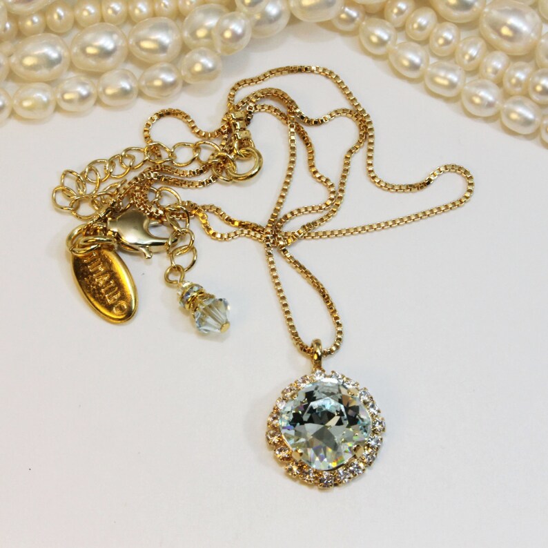 May include: A gold chain necklace featuring a large, round, clear crystal pendant encircled by smaller crystals. The pendant is secured to a gold chain with a lobster clasp. The chain also includes a small, clear crystal charm.