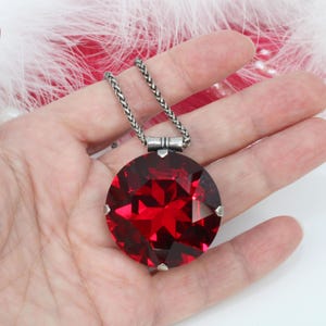 May include: A large, round, faceted red pendant necklace with a silver chain. The pendant is a deep ruby color and has a geometric design. The chain is a woven style. The necklace is a statement piece.