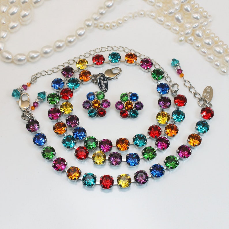 Puede incluir: Un conjunto de collar y pendientes de cristal arco&iacute;ris. El collar est&aacute; hecho de tres hileras de cadena plateada con cuentas de cristal de colores arco&iacute;ris. Los pendientes son peque&ntilde;os tachuelas en forma de flor con cristales de colores arco&iacute;ris.