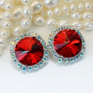 May include: Two red, round earrings with a silver-colored, iridescent crystal border. The earrings have a large, faceted, ruby-red center stone. The background features a strand of white pearls.