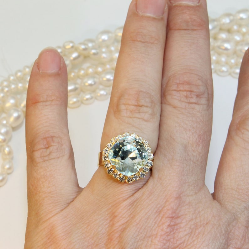 May include: A gold ring featuring a large, light blue gemstone encircled by smaller clear gemstones.