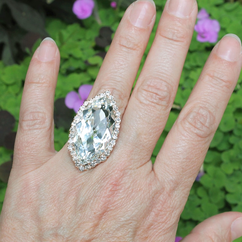 May include: A large, clear, marquise-shaped ring with a diamond-like stone surrounded by smaller diamonds. The ring is on a person's finger.