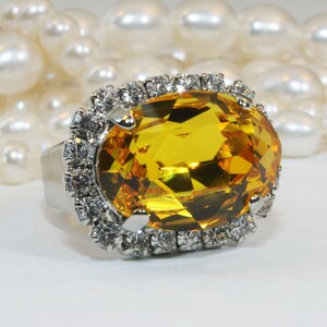 May include: A silver-toned ring featuring a large, oval-cut, golden-yellow gemstone at the center. The central stone is surrounded by a halo of small, clear, round-cut white crystals set in a silver-colored metal frame with a textured band.