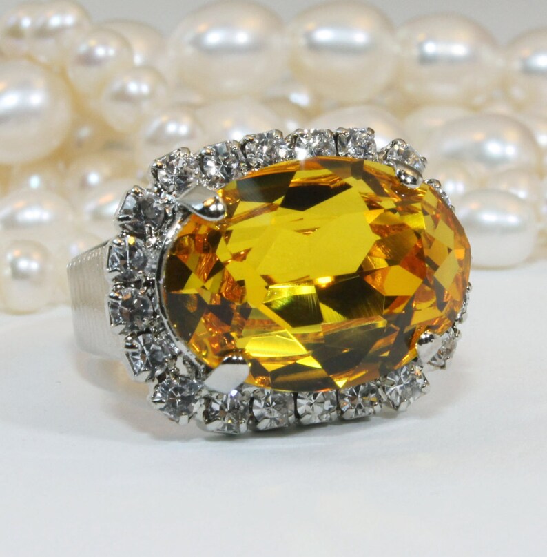 May include: A silver-toned ring featuring a large, oval-cut, golden-yellow gemstone at the center. The central stone is surrounded by a halo of small, clear, round-cut white crystals set in a silver-colored metal frame with a textured band.